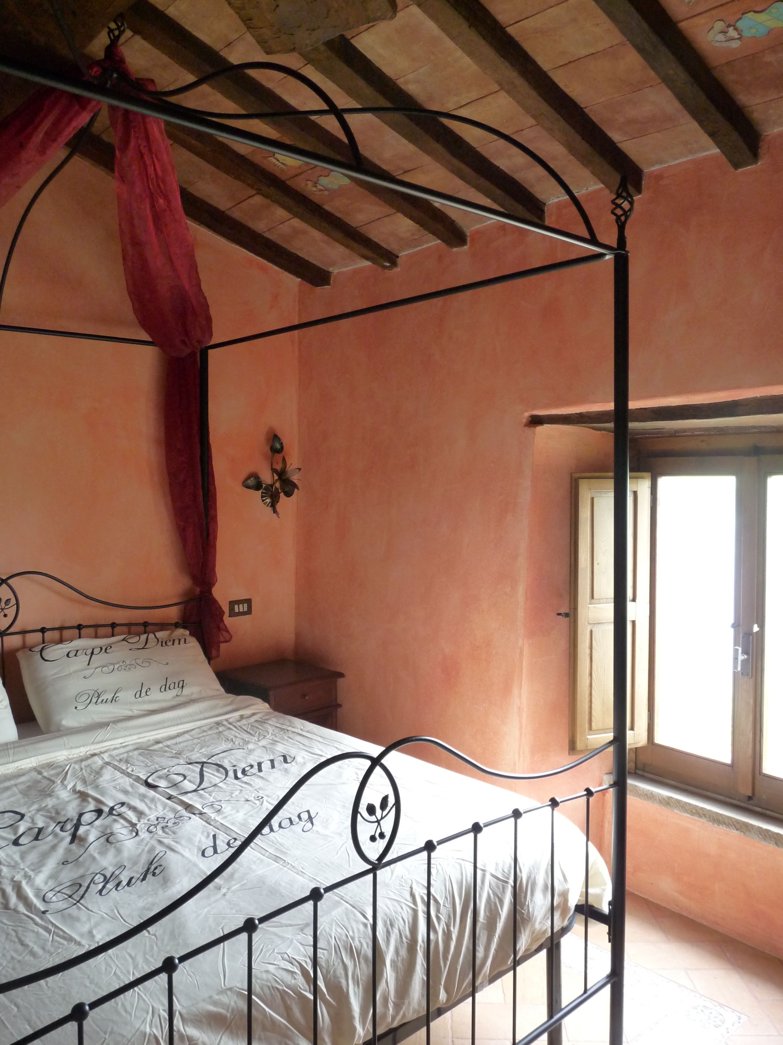 Una cama con dosel y cortinas, una mesita de noche, una lámpara de pared y una ventana con contraventanas de madera.