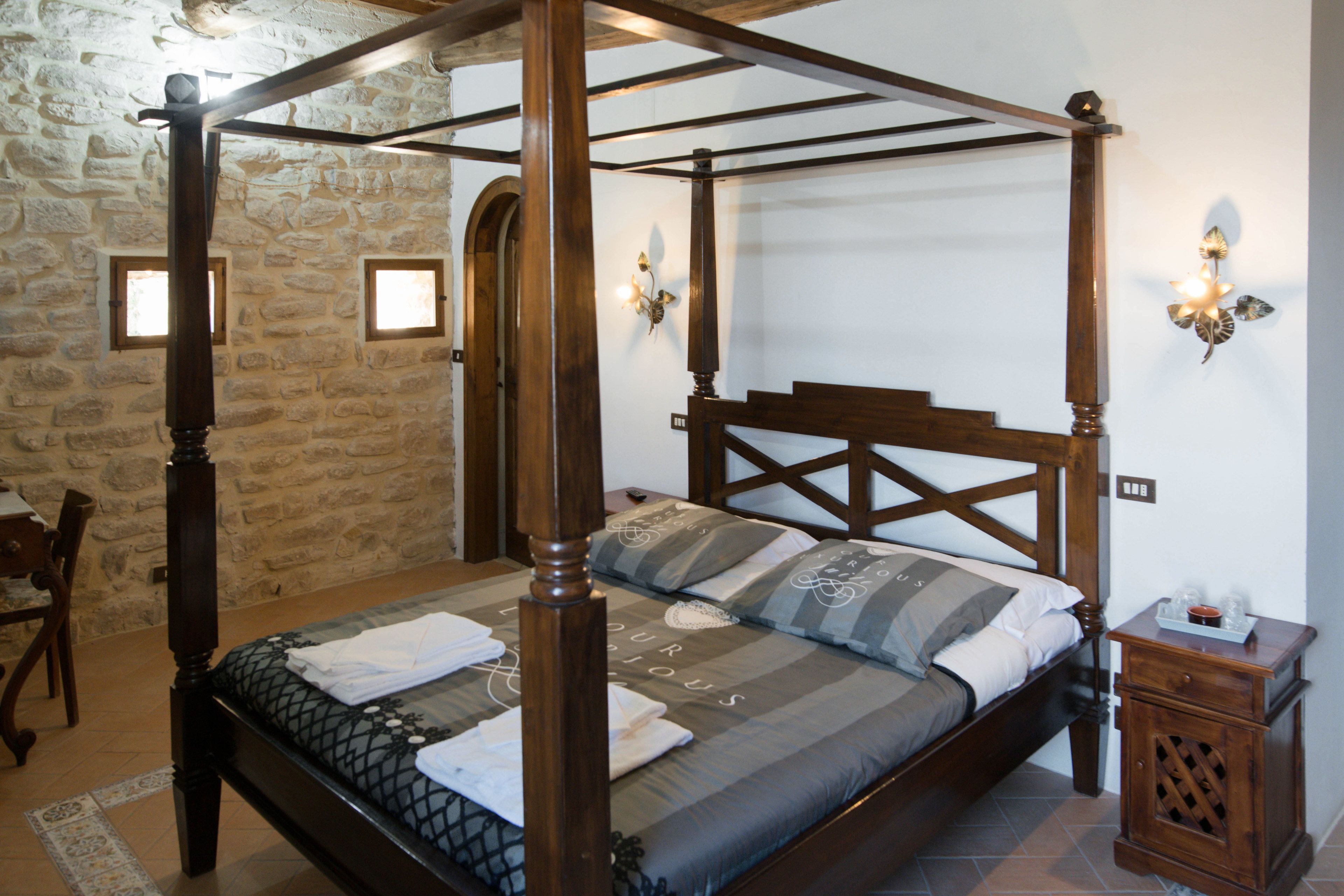 Una cama con dosel de madera oscura y una colcha gris, una mesita de noche de madera con una lámpara, y una pared de piedra con una puerta al fondo.
