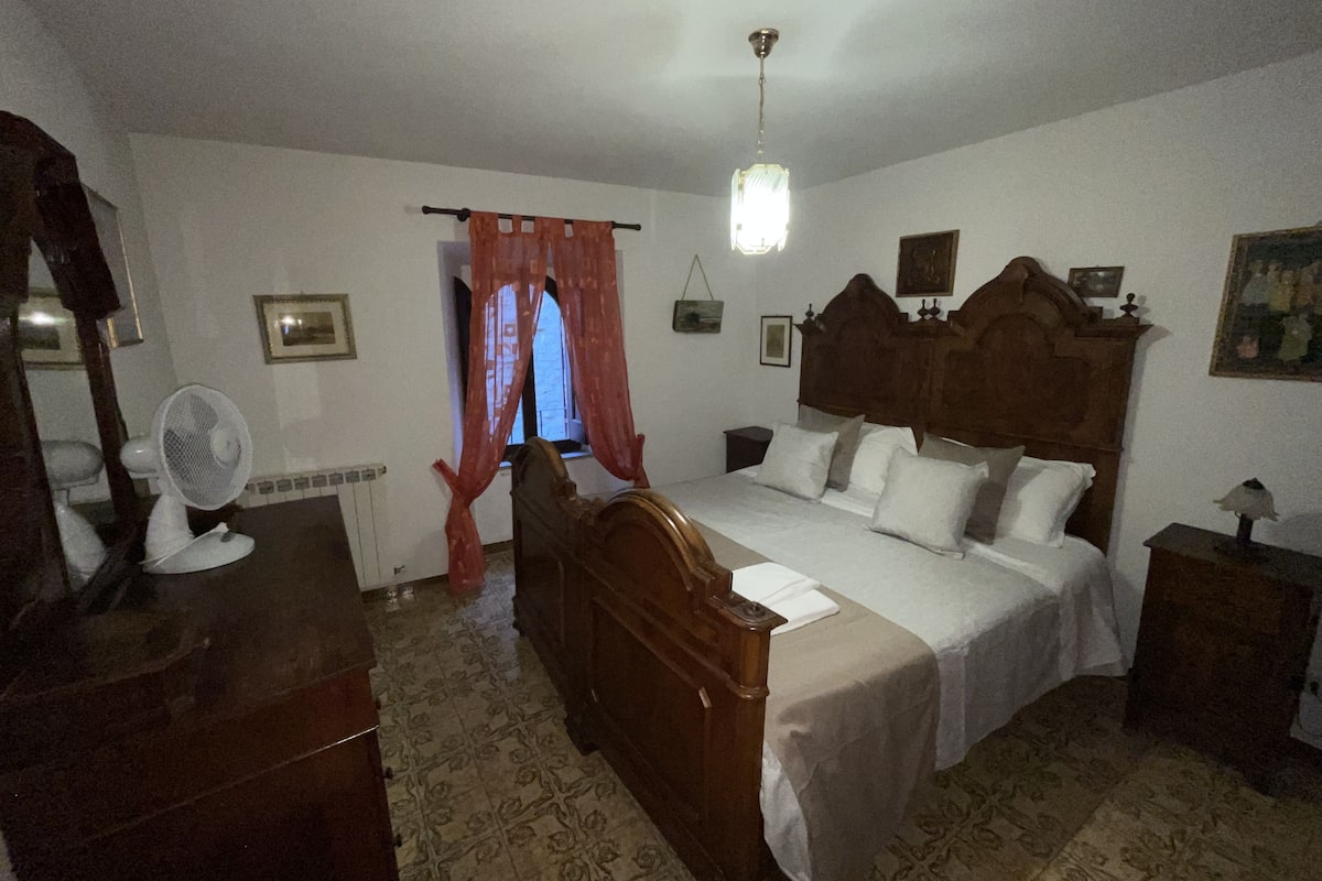 A bedroom with a wooden bed, a bedside table, a nightstand, a mirror, a fan, a window with curtains, and a hanging light fixture.
