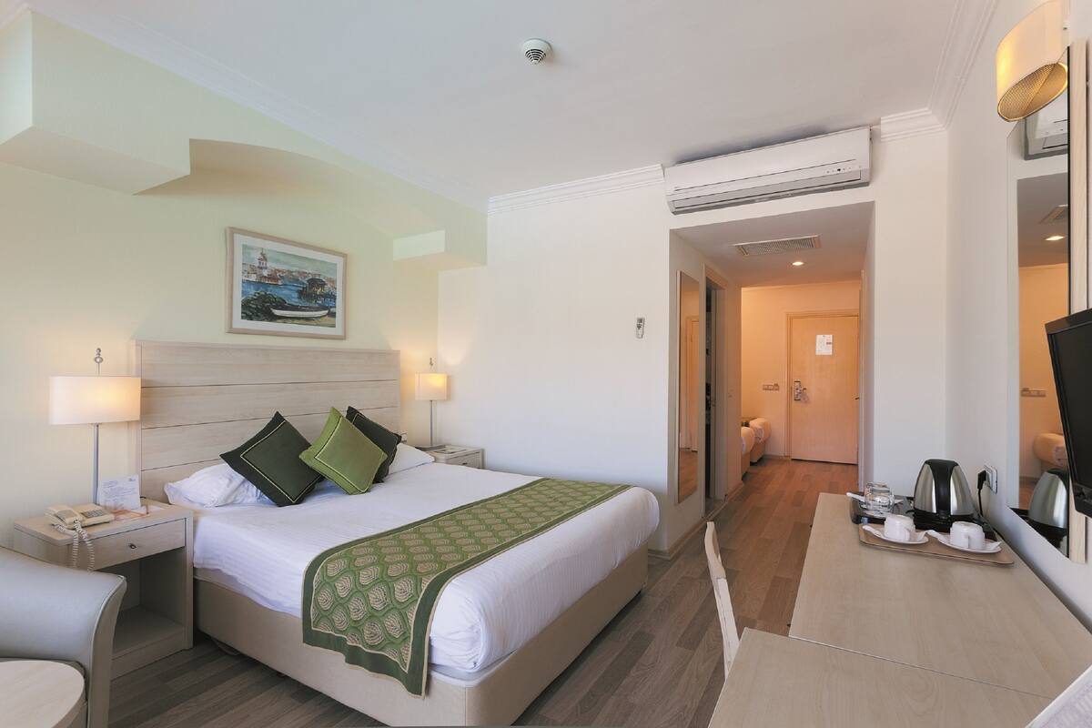 Habitación de hotel con cama, mesitas de noche, un escritorio con bandeja de tazas y tetera, un cuadro en la pared y un equipo de aire acondicionado.