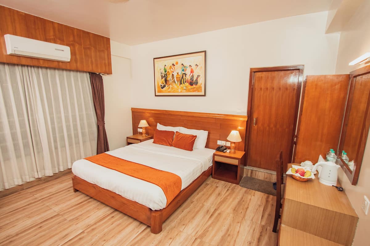 A hotel room with a wooden bed, a painting on the wall, a wooden wardrobe, and a bedside table with a lamp and a fruit basket.