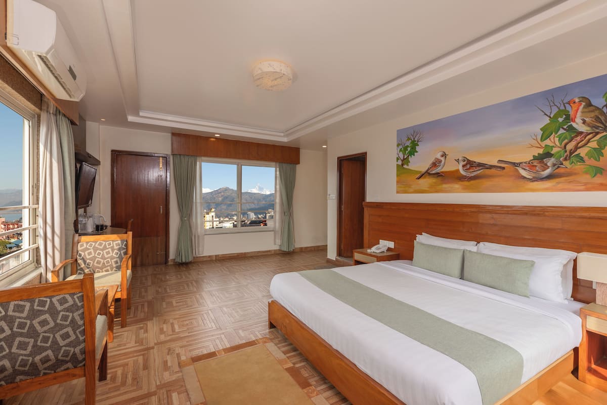 A hotel room with a large bed, a dining area, a painting of birds, and a view of the city.