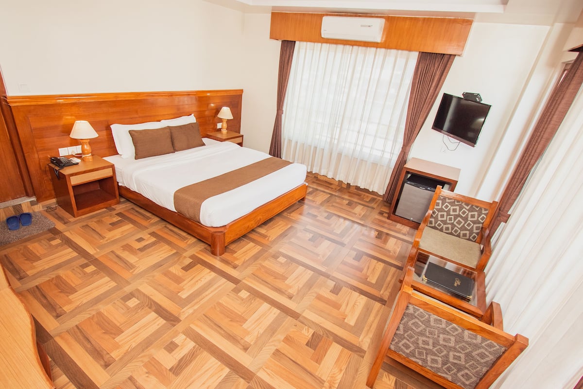A hotel room with a wooden floor, a bed with a headboard, a nightstand, a lamp, a TV, and a small table with a chair.