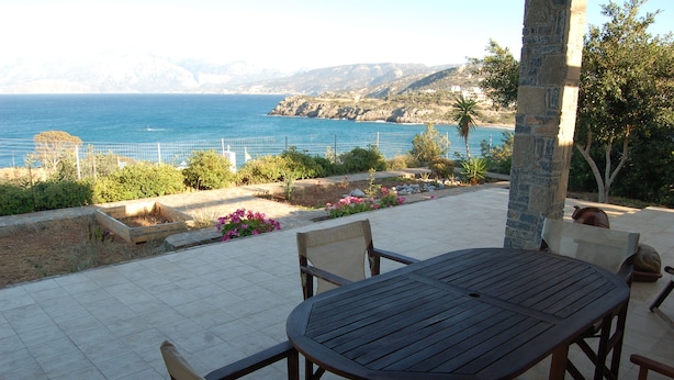 Eine Terrasse mit einem runden Tisch und Stühlen, mit Blick auf eine Küstenlandschaft und dem Meer sowie ferne Berge.