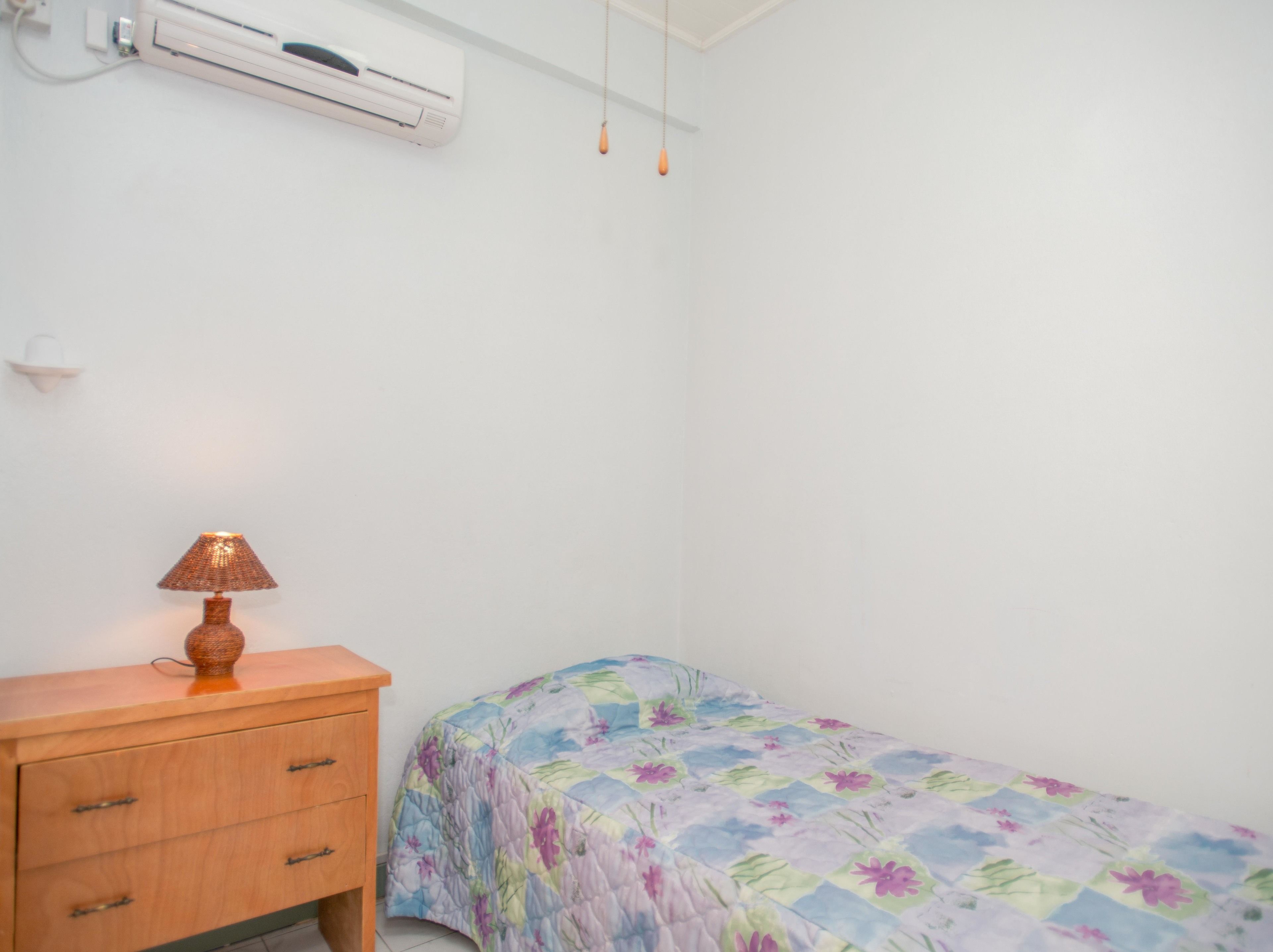 A small, single-bed room with a wooden dresser, a floral bedspread, and a wall-mounted air conditioner.