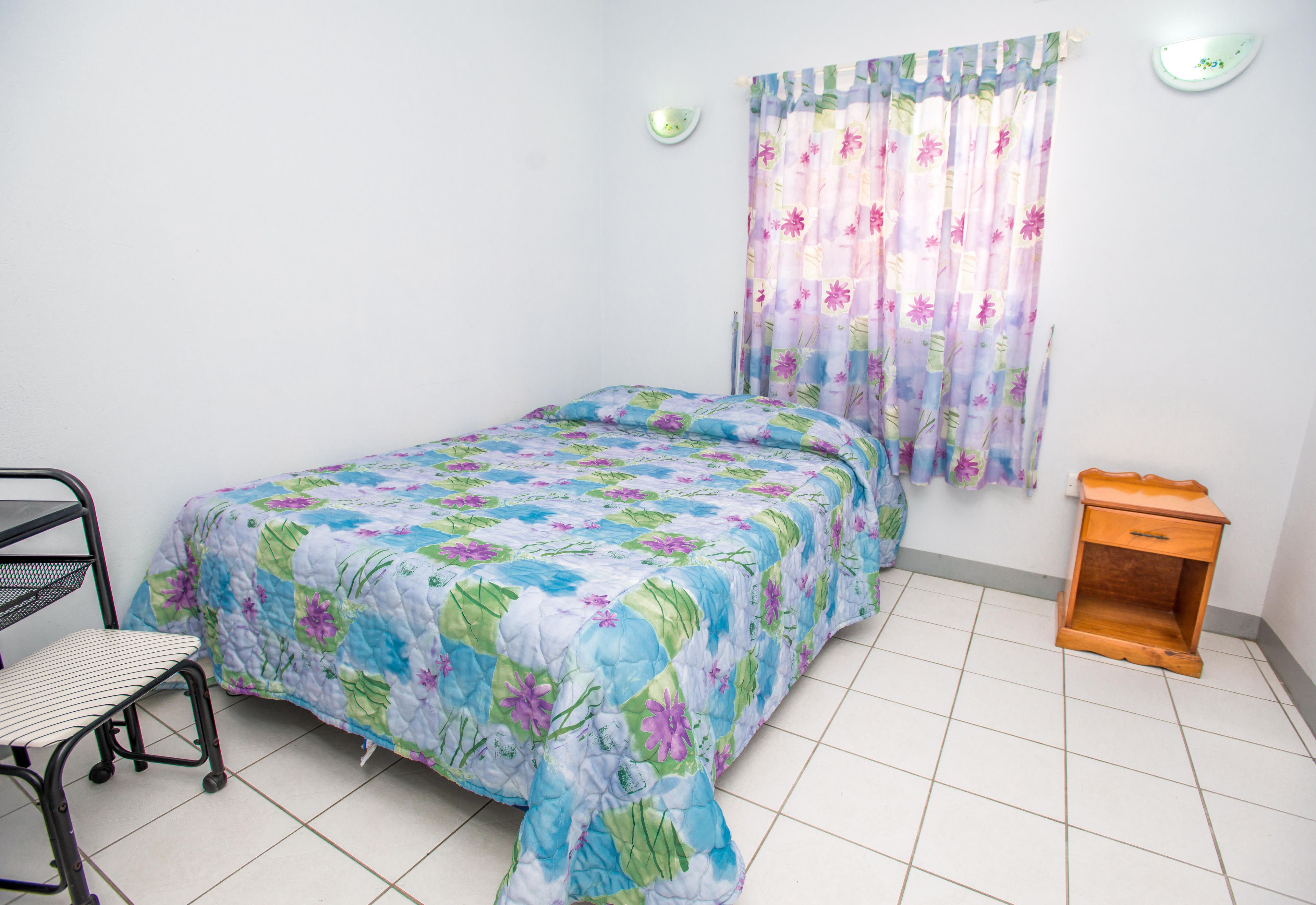 Un dormitorio con cama, una silla, una mesita de noche y una ventana con cortinas.