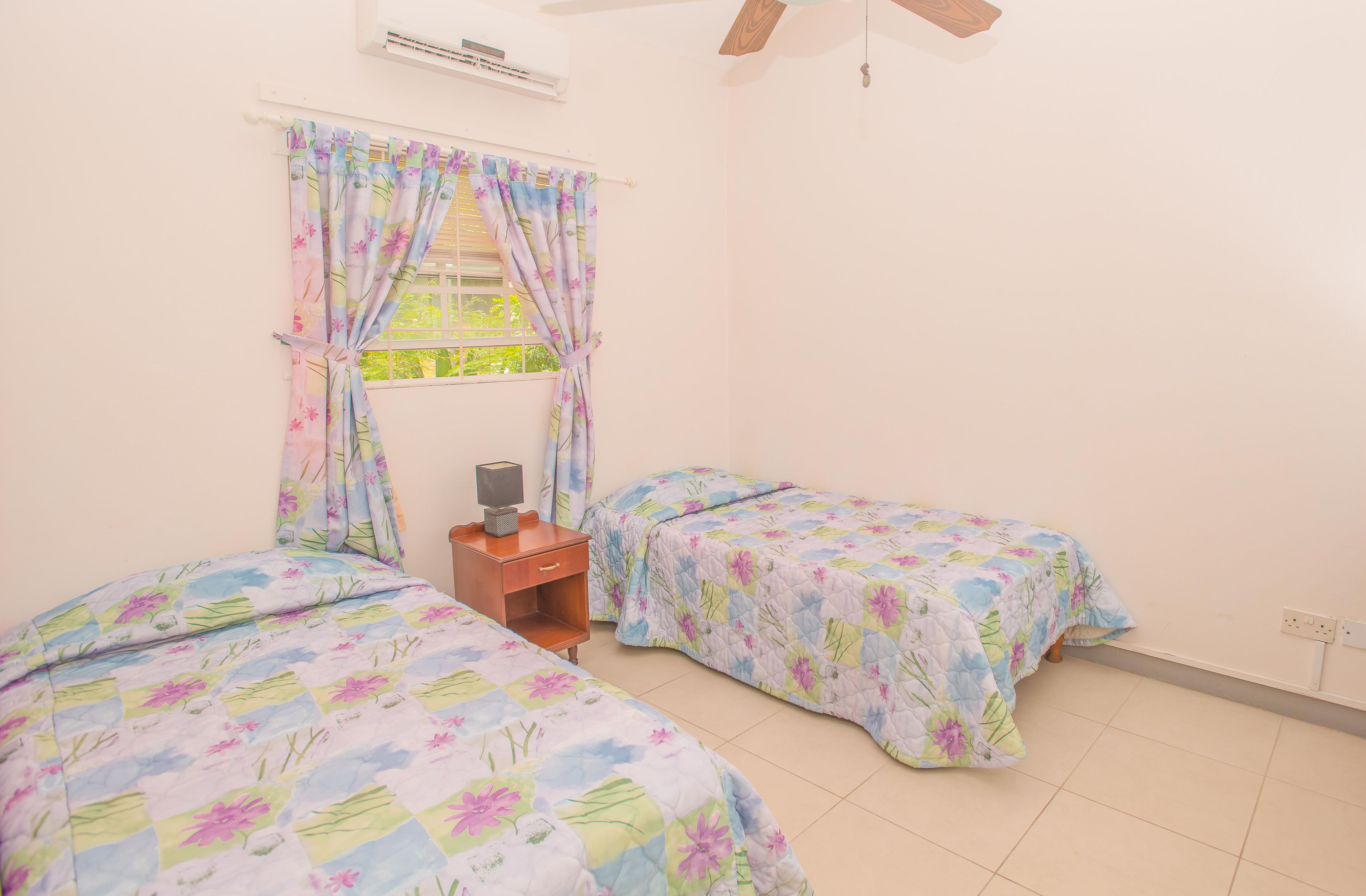 Quarto com duas camas, mesa de cabeceira, ar-condicionado e janela com cortinas florais.