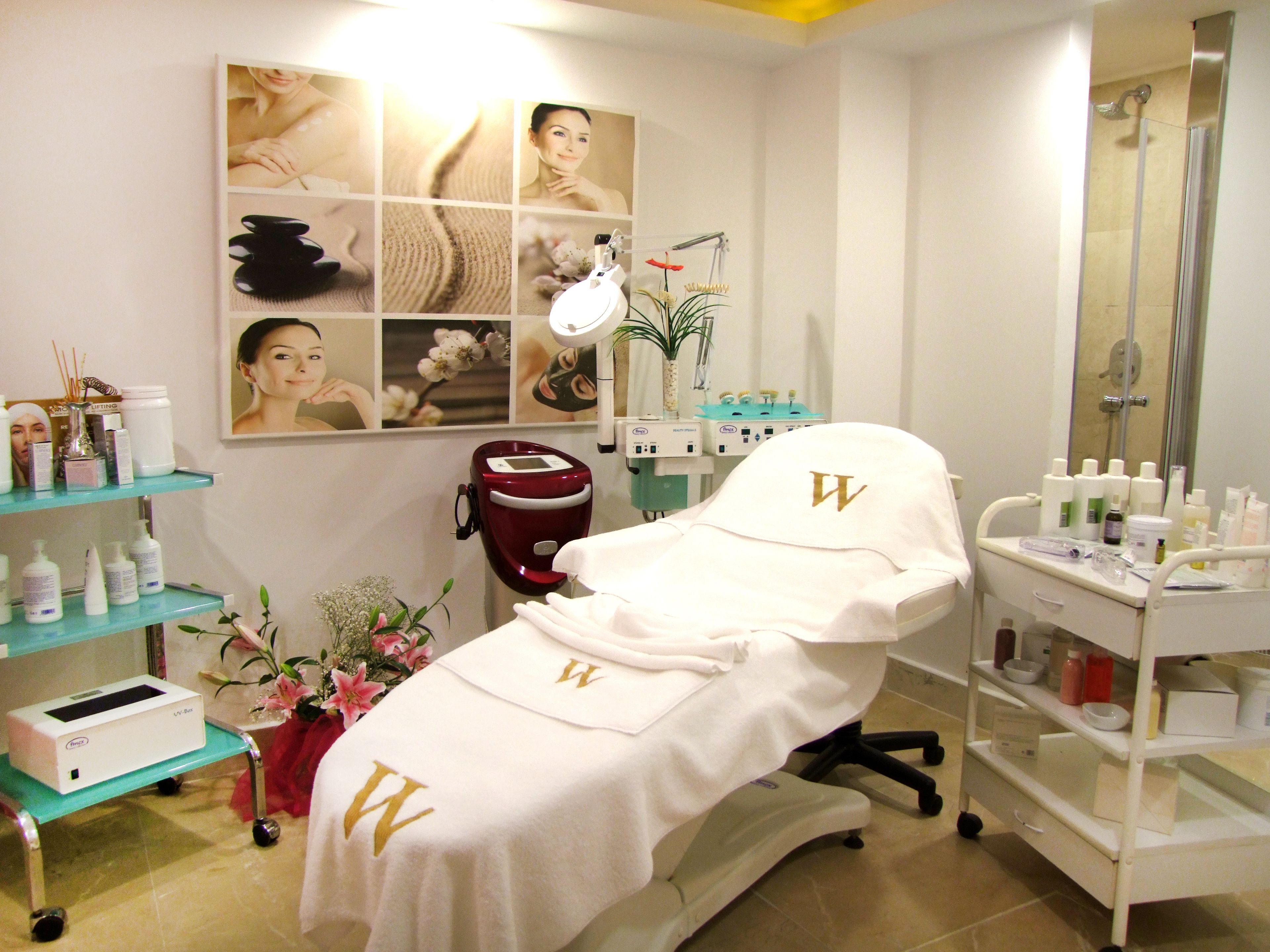 A medical or spa treatment room with a treatment bed, shelves with various products, a red massage chair, and a wall with framed photos.