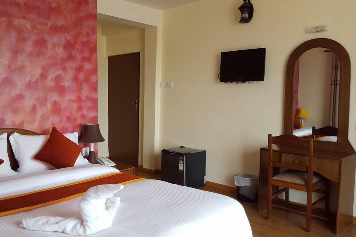 Habitación de hotel con cama, escritorio, silla, televisor, espejo y cubo de basura.