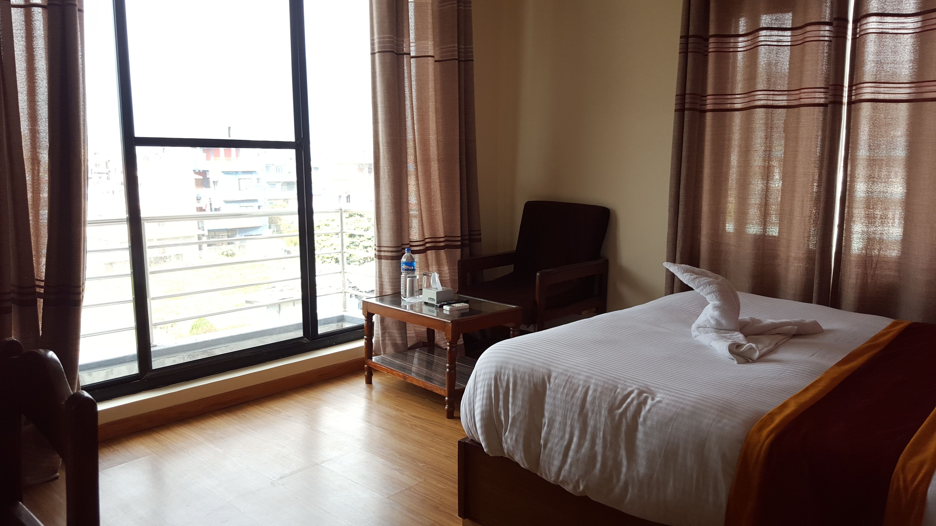 A hotel room with a large window, a bed with a white comforter, a wooden nightstand, a chair, and a view of the outside.