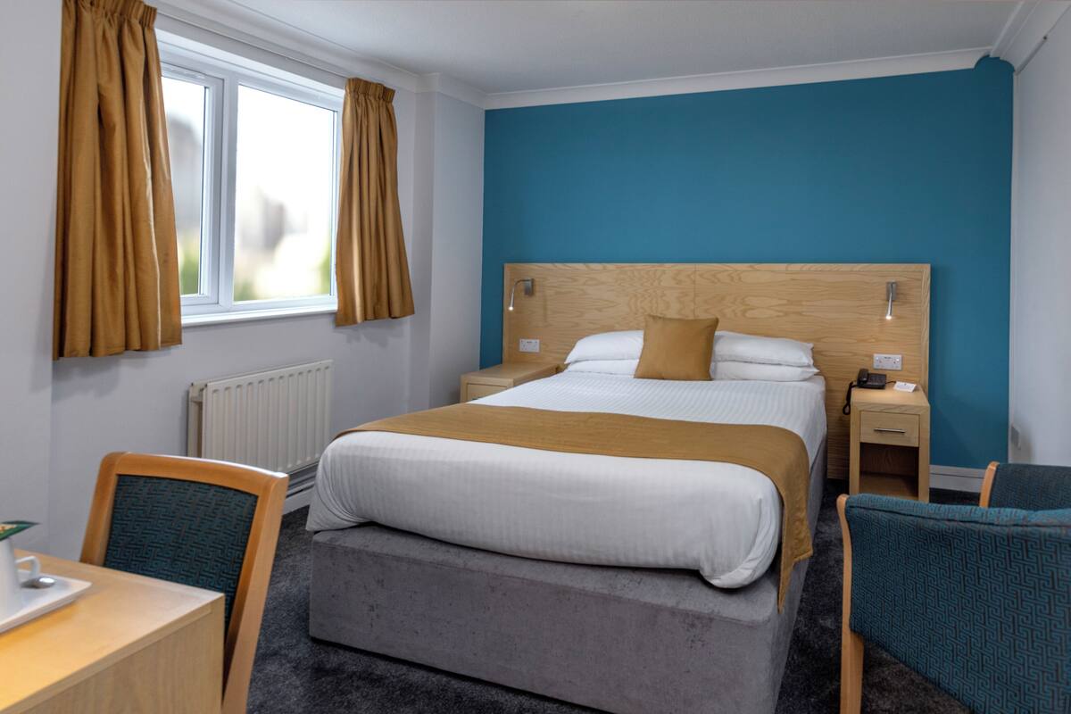 A hotel room with a bed, bedside table, chair, and a window with curtains.