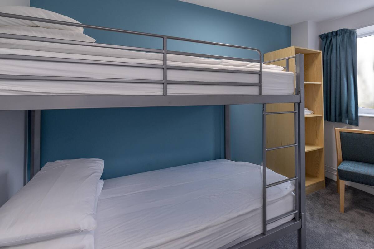 A bunk bed with white bedding, a desk with a chair, and a bookshelf.