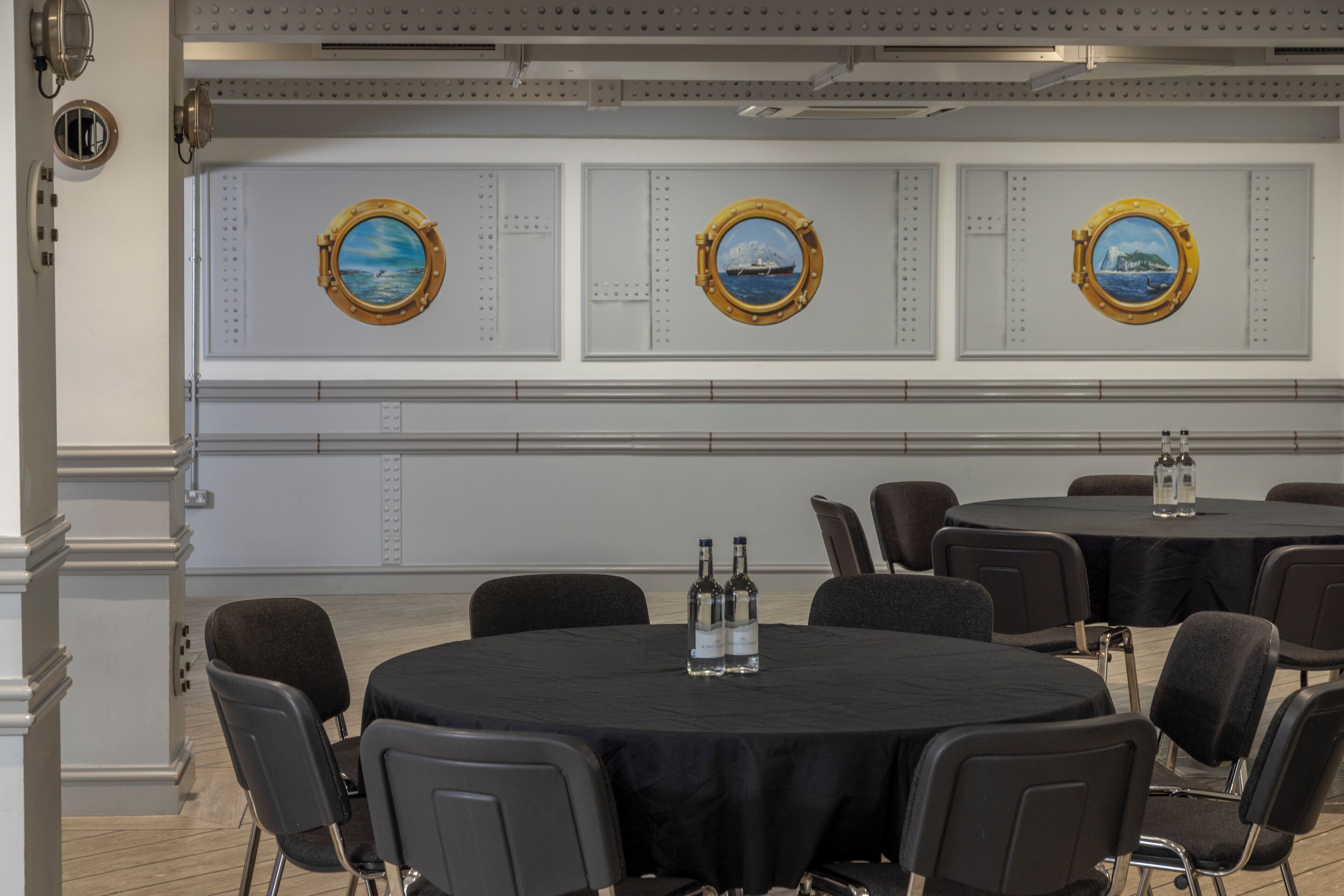 A conference room with round tables, black tablecloths, and chairs. There are three wall-mounted porthole-style windows with maritime scenes.