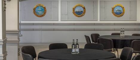 A conference room with round tables, black tablecloths, and chairs. There are three wall-mounted porthole-style windows with maritime scenes.