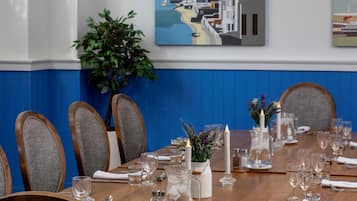 Uma sala de jantar com uma mesa de madeira redonda posta para seis pessoas, rodeada por cadeiras estofadas cinzentas. Há luminárias pendentes sobre a mesa, e uma obra de arte emoldurada retratando uma paisagem costeira é visível na parede.