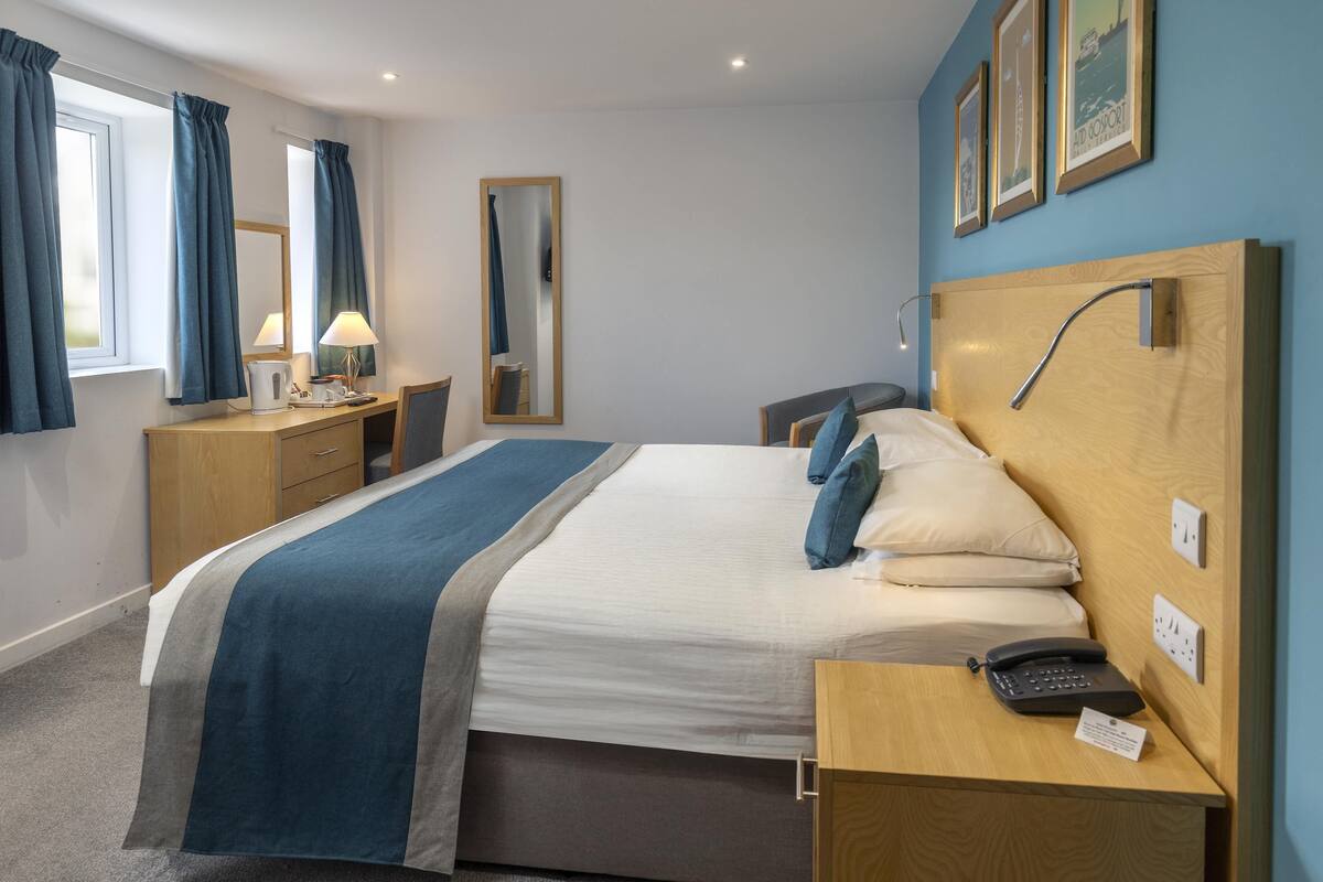 A hotel room with a bed, bedside table, lamp, mirror, and a wall with framed pictures.