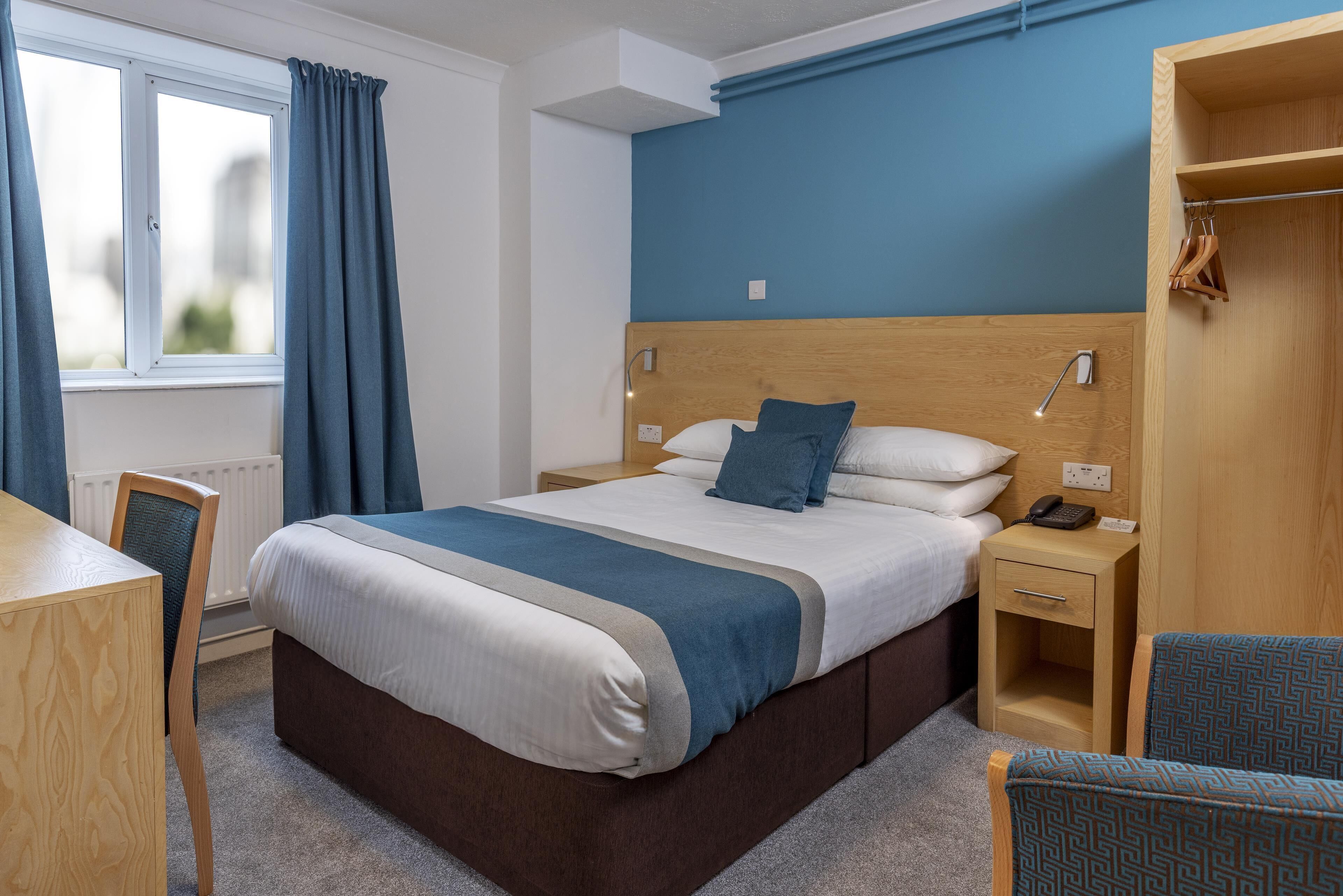 A hotel room with a bed, bedside table, chair, wardrobe, and a window with curtains.