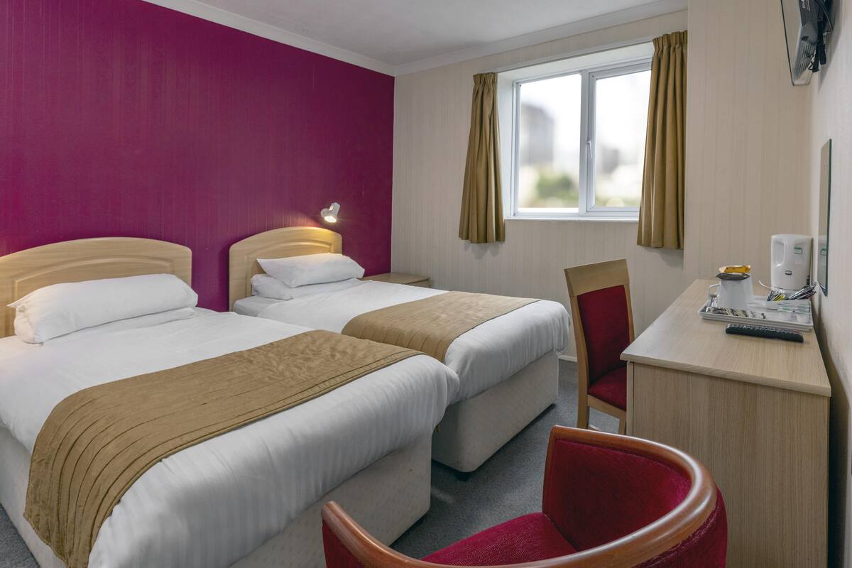 A hotel room with two beds, a desk with a chair, a window with curtains, and a wall-mounted TV.