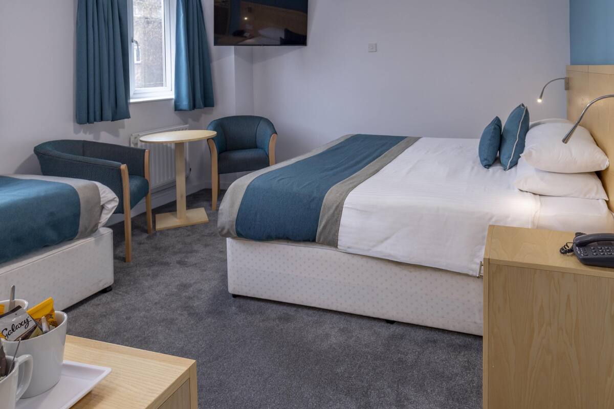A hotel room with two beds, a small table, a chair, a bedside table with a phone, and a TV mounted on the wall.