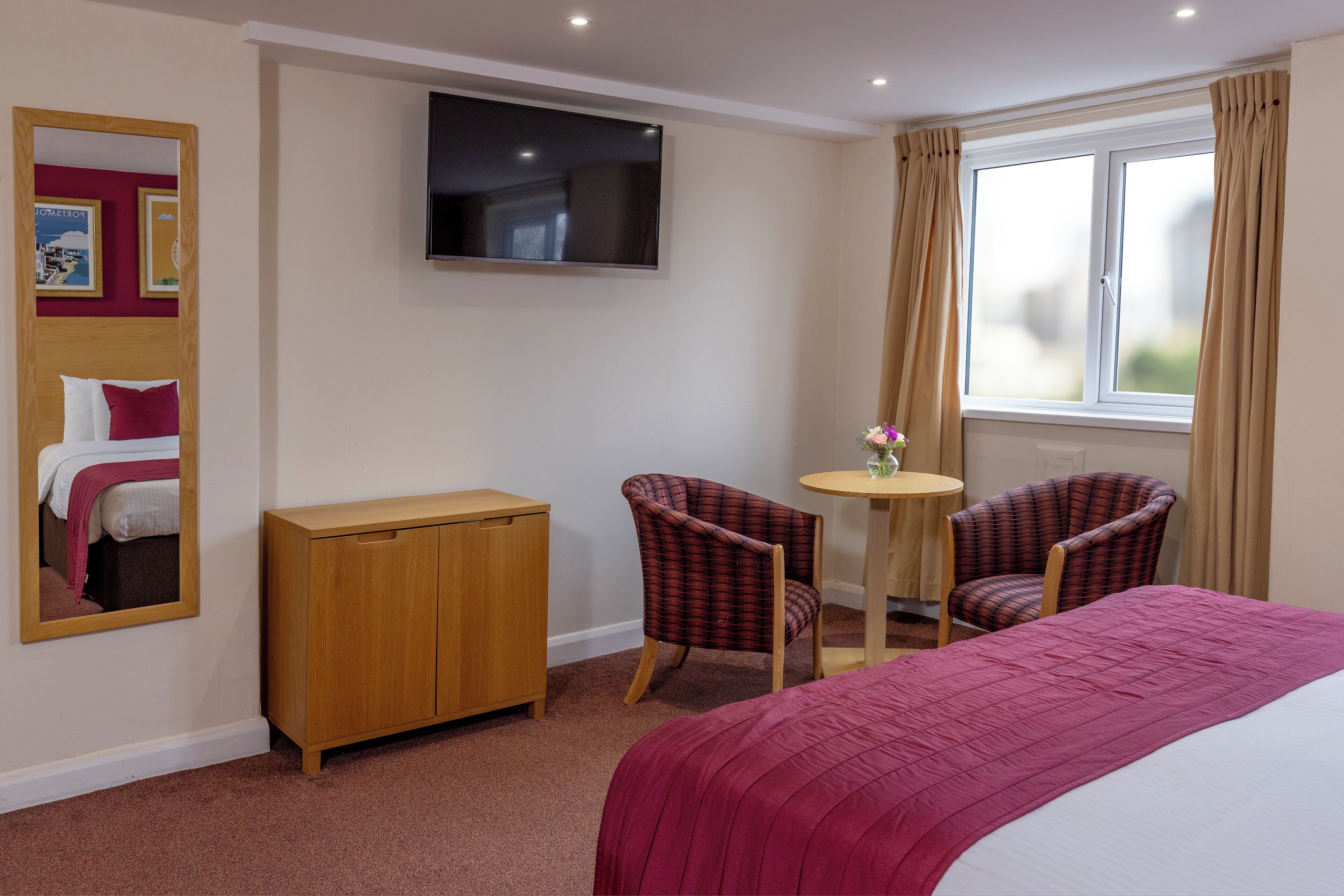 A hotel room with a bed, a TV, a wooden cabinet, two chairs, a small table, and a window with curtains.
