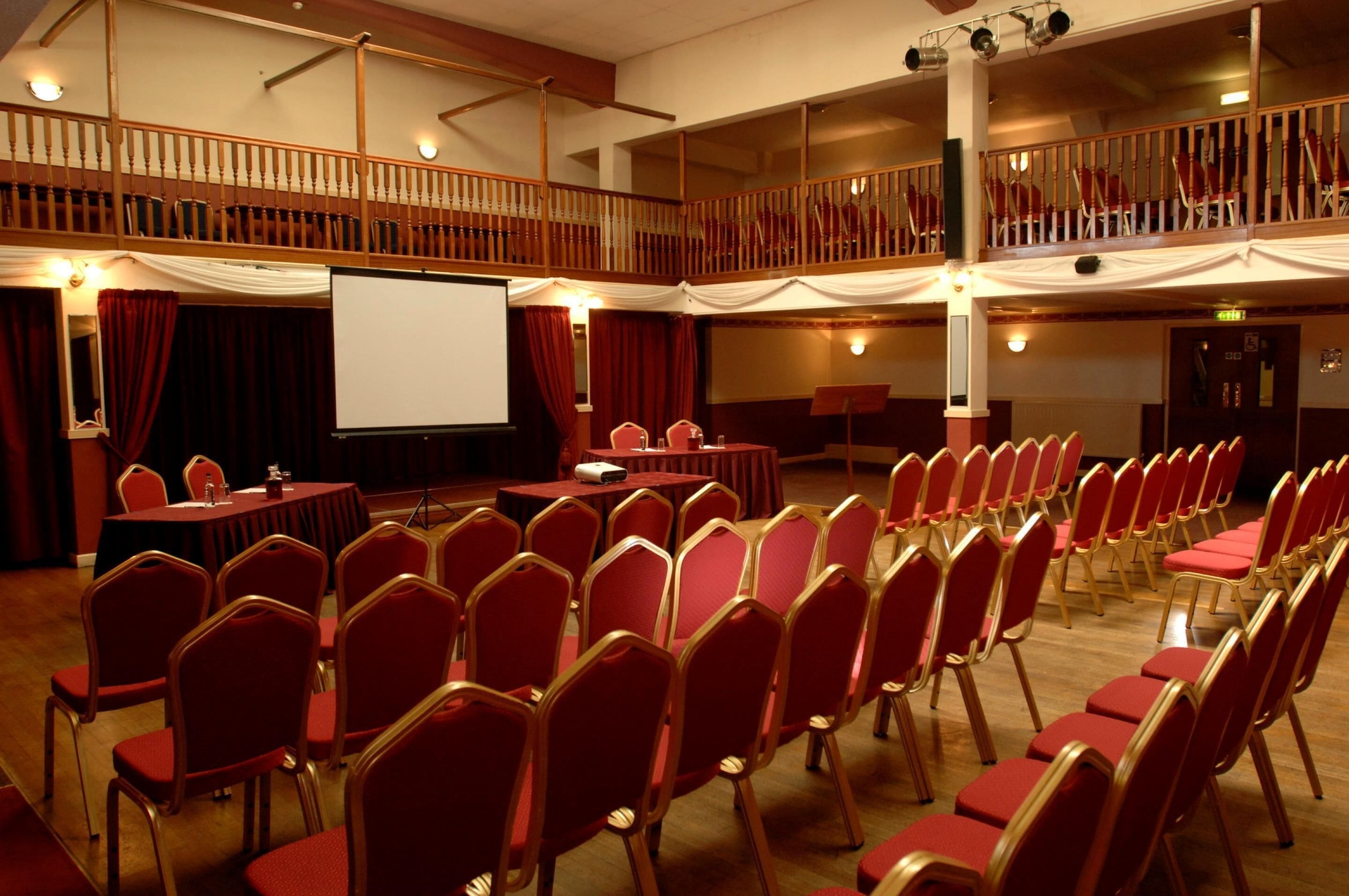 Un salón de conferencias con asientos escalonados, un escenario con pantalla y un balcón.
