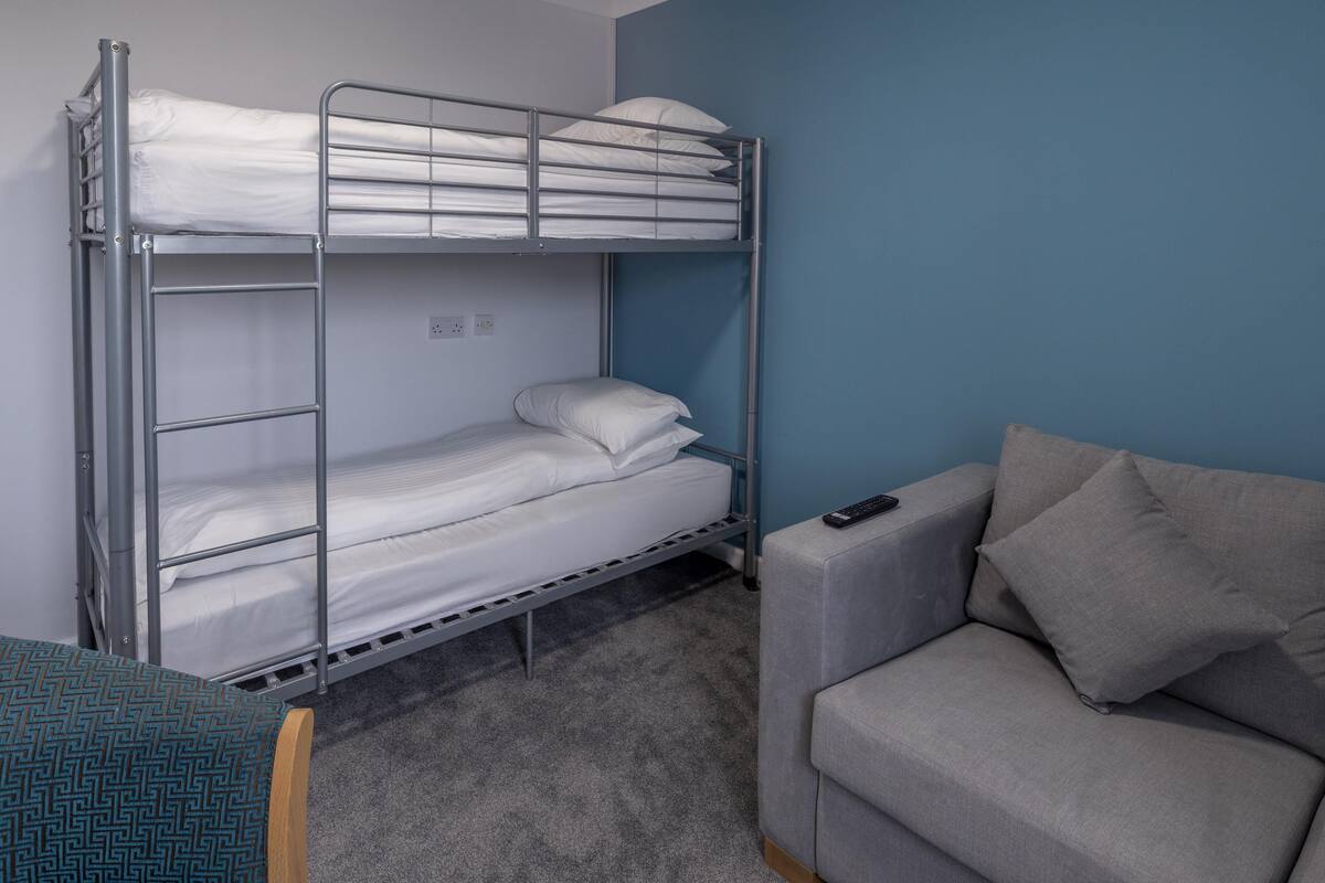 A bunk bed with white bedding, a grey sofa with cushions, and a patterned chair.
