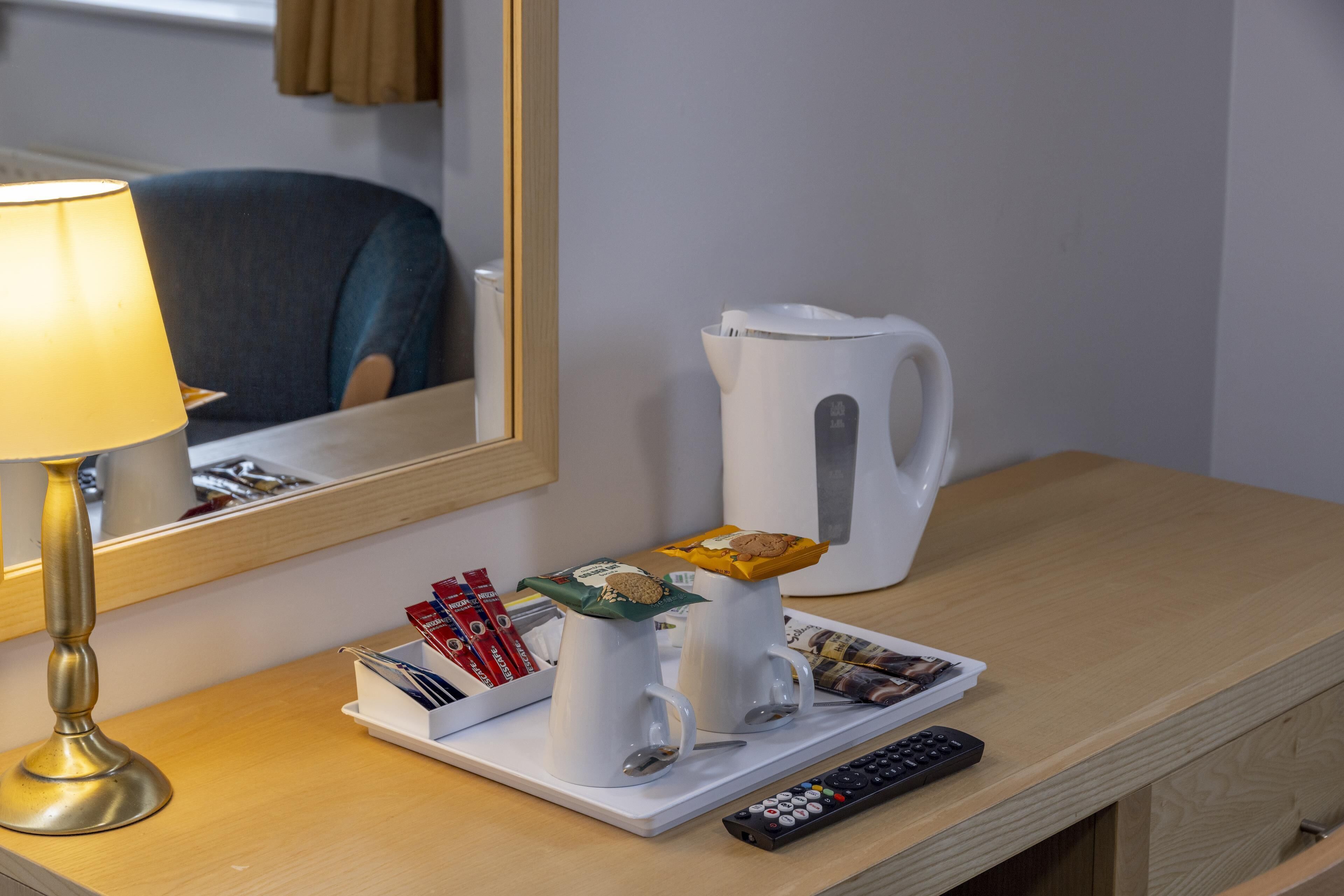 Ett hotellrum med ett skrivbord i trä, en lampa, två vita muggar med snacks, en fjärrkontroll och en vit kanna.