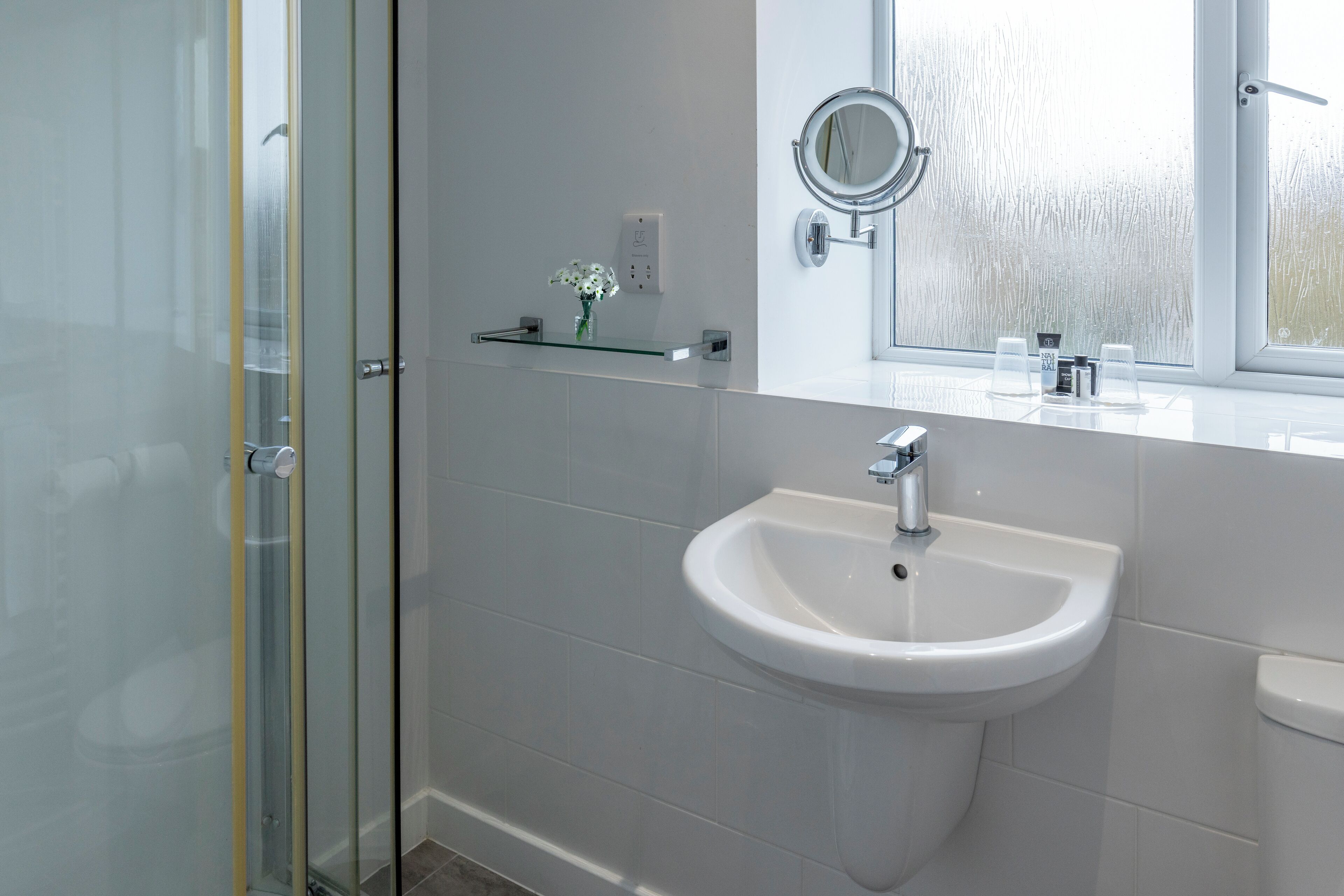 A modern bathroom with a glass shower enclosure, a white sink with a mirror, and a window with frosted glass.