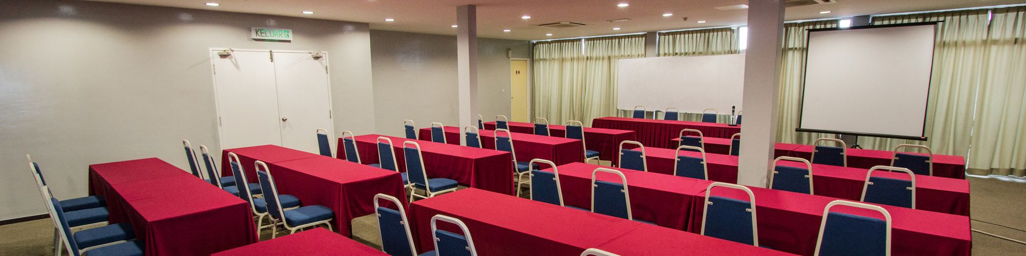 A conference room with rows of tables and chairs, a projector screen, and a door labeled "KELUARAN".