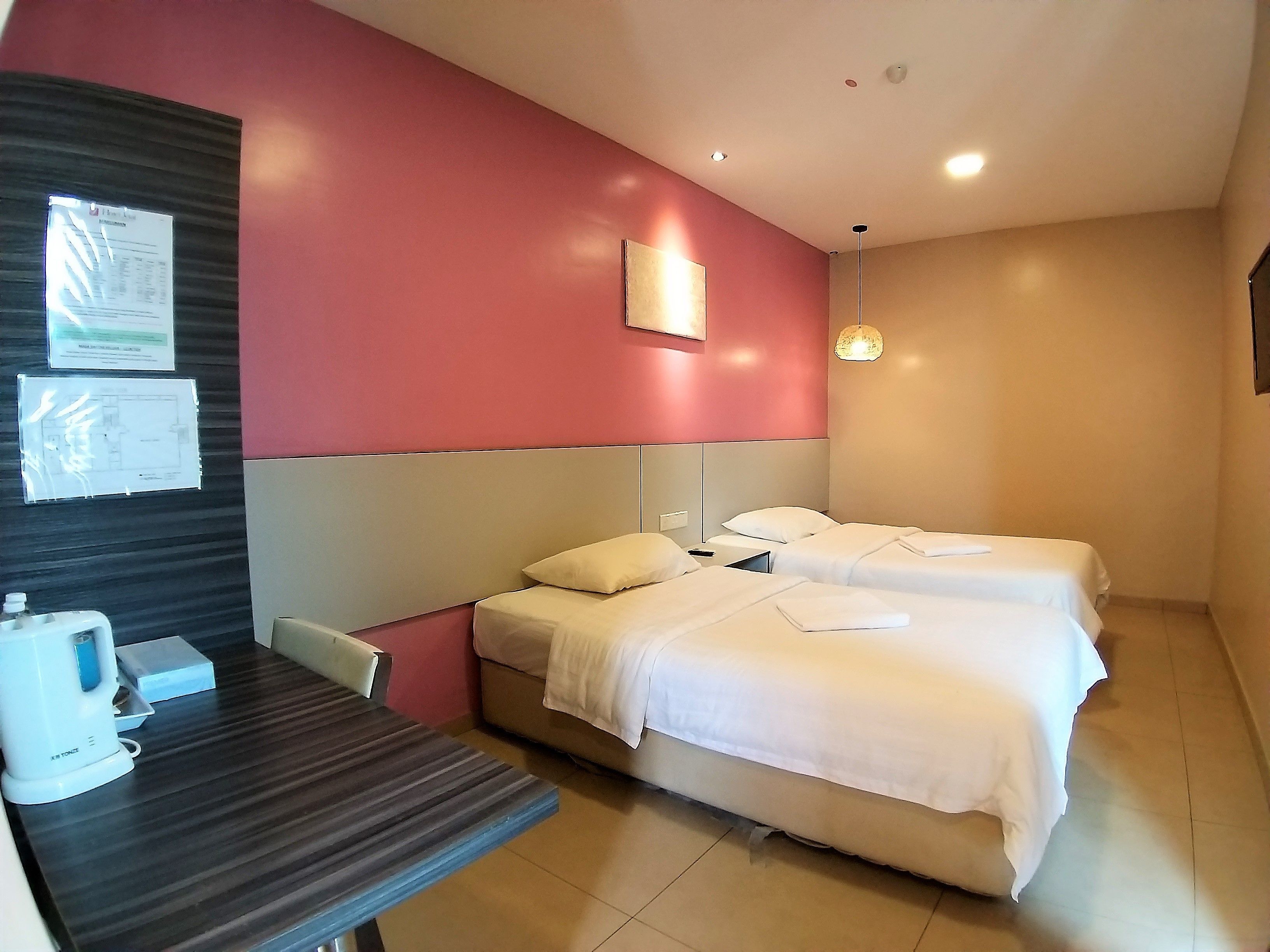 A hotel room with two beds, a desk, a chair, a coffee maker, and a wall-mounted light fixture.