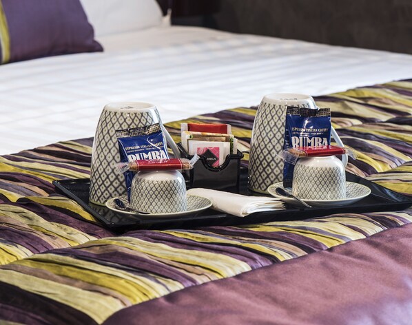 A tray with two cups of espresso, a small bag of biscotti, and a container of biscotti on a bed with a patterned bedspread.