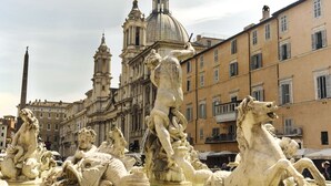 Una fuente clásica con múltiples estatuas, rodeada de edificios históricos y una iglesia destacada al fondo.