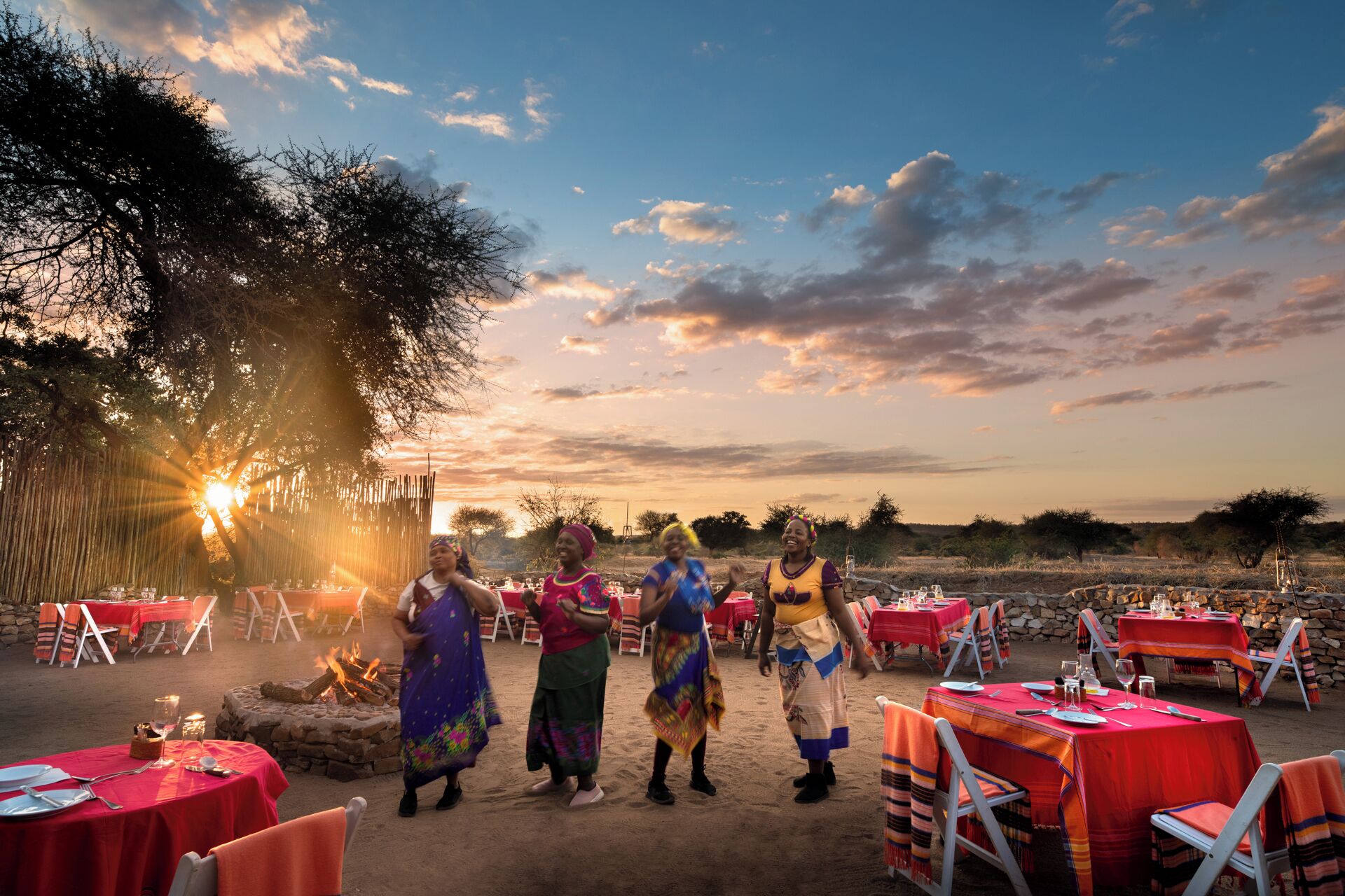 An outdoor dining area with tables set for a meal, surrounded by natural scenery and a sunset in the background.
