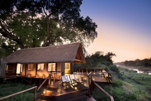 A rustic wooden cabin with a thatched roof, a wooden deck, and a river view. - Pafuri Camp by RETURN Africa (Mutale)