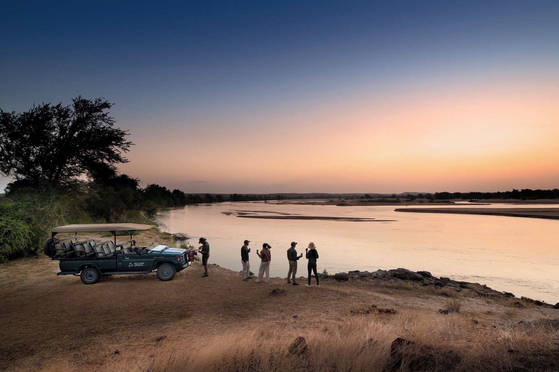 A group of people standing by a river at sunset, with a safari vehicle parked nearby.