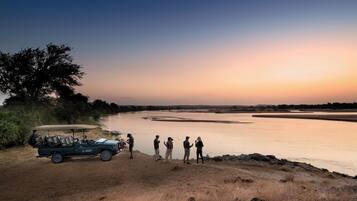 A group of people standing by a river at sunset, with a safari vehicle parked nearby.