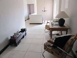 A room with a wheelchair, a small table with a lamp, a bookshelf, and a bed in the background. - Karoo Sun Boutique Guest House (Oudtshoorn)
