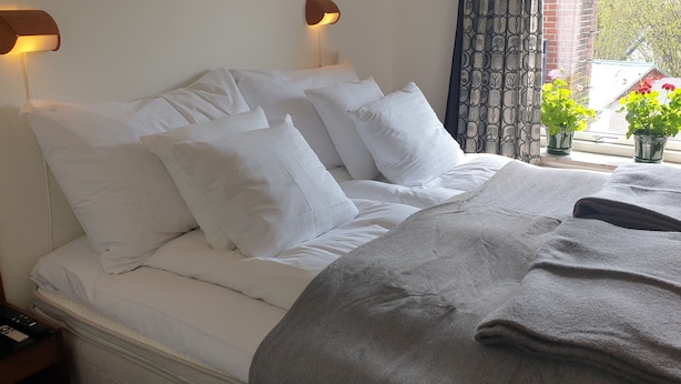 Una cama bien tendida con sábanas blancas y una manta gris, una lámpara de pared y una ventana con vistas a follaje y un muro de ladrillo.