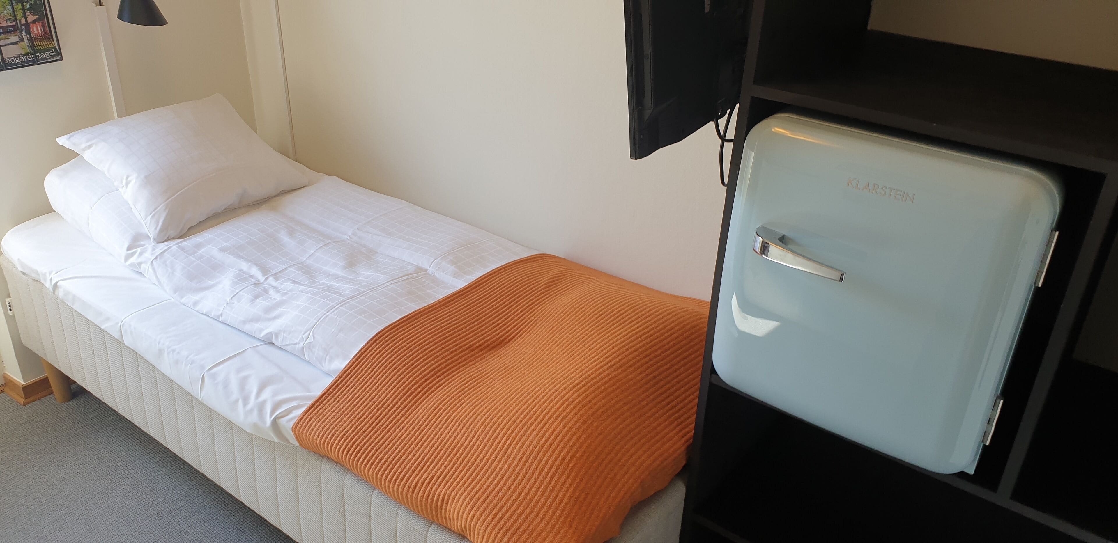 A single bed with white bedding and an orange throw blanket, a small black TV, and a light blue refrigerator.
