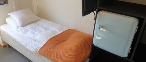 A single bed with white bedding and an orange throw blanket, a small black TV, and a light blue refrigerator.