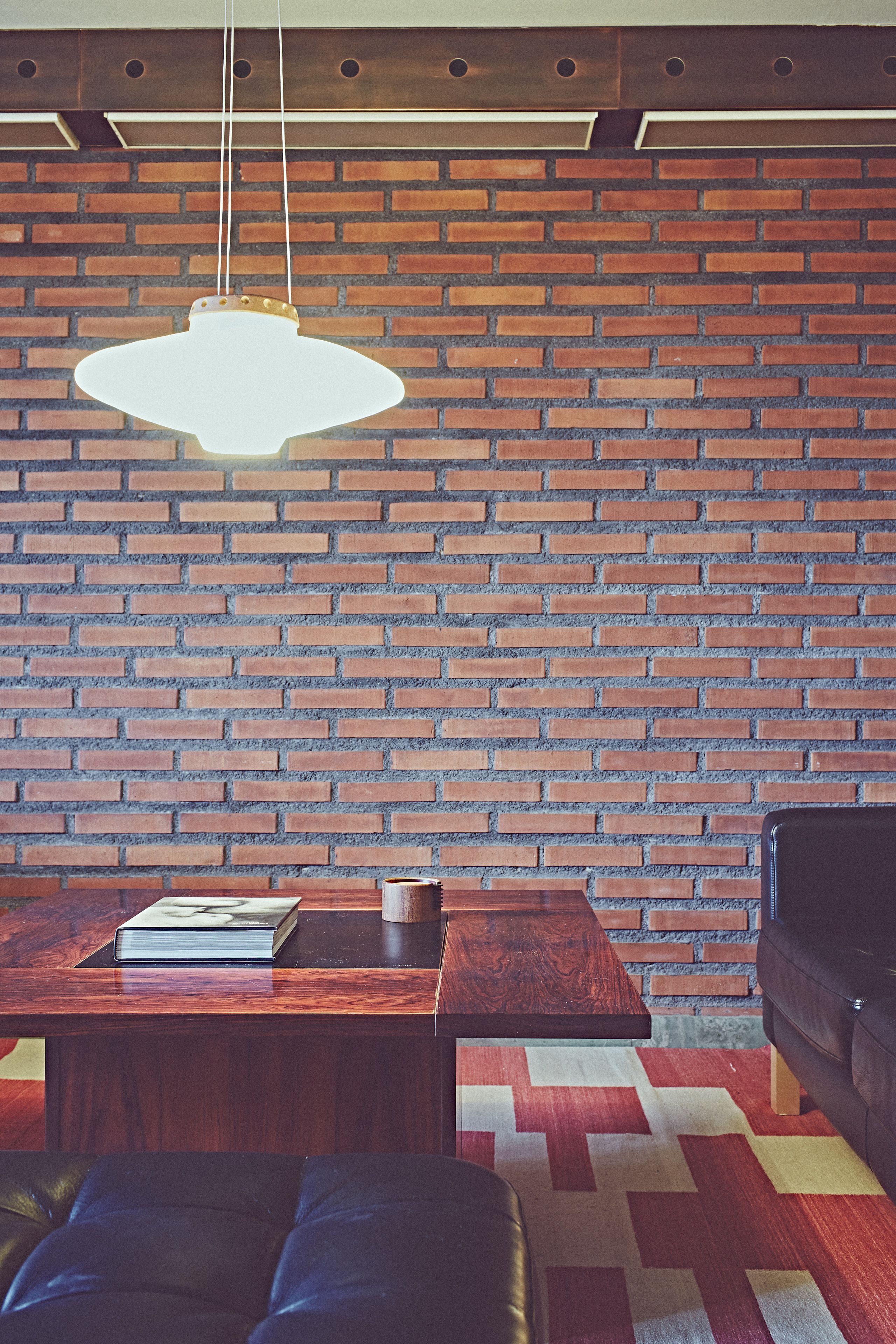 A room with a brick wall, a hanging light fixture, a wooden table with a book and a cup, a leather couch, and a patterned rug.