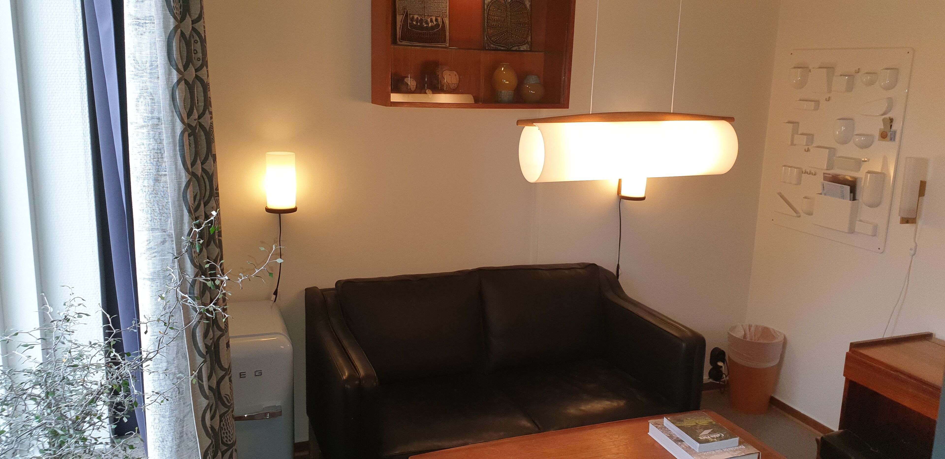 A room with a black leather sofa, a wooden table with a book, a lamp, a small trash bin, and a wall-mounted light switch panel.