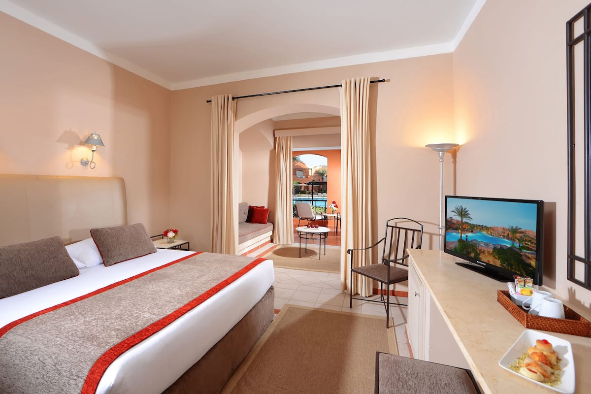 A hotel room with a bed, a desk with a TV, a chair, and a view of an outdoor pool area.