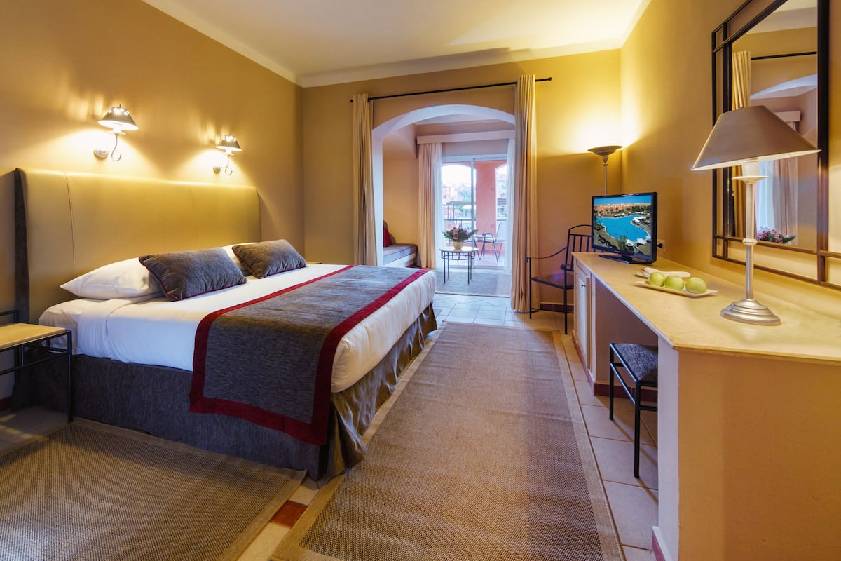 A hotel room with a bed, a desk with a TV, and a view of the outside.