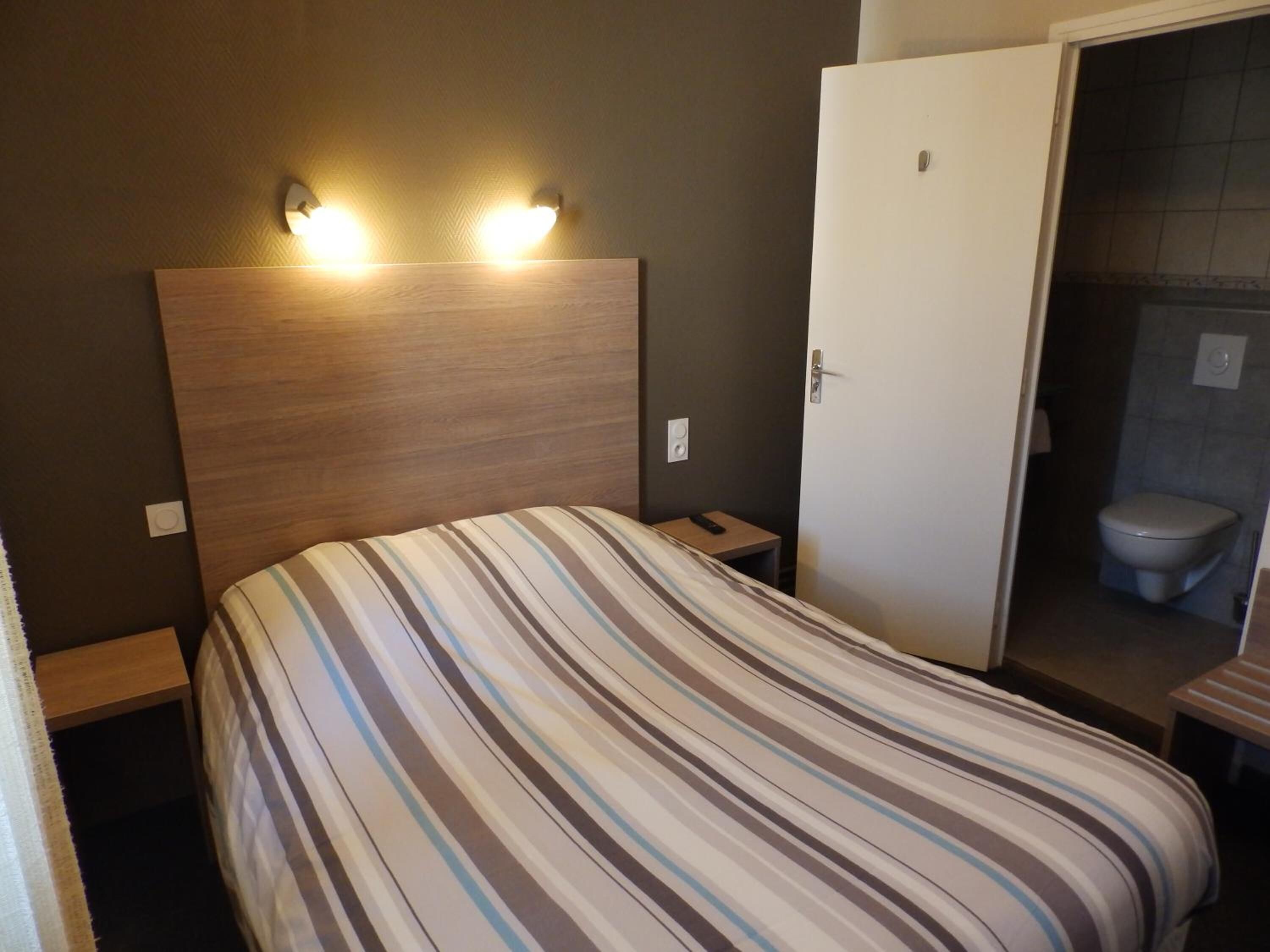 Habitación de hotel con cubrecama a rayas, cabecera de madera, mesitas de noche y baño con inodoro y lavamanos.
