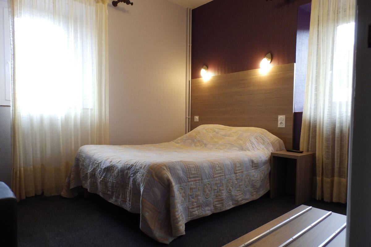A single bed in a room with a wooden headboard, a bedside table, and a window with sheer curtains.