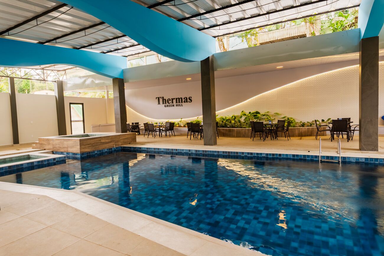 An indoor swimming pool with a lounge area, a sign reading 'Thermas GREEN HILL,' and a view of greenery outside.