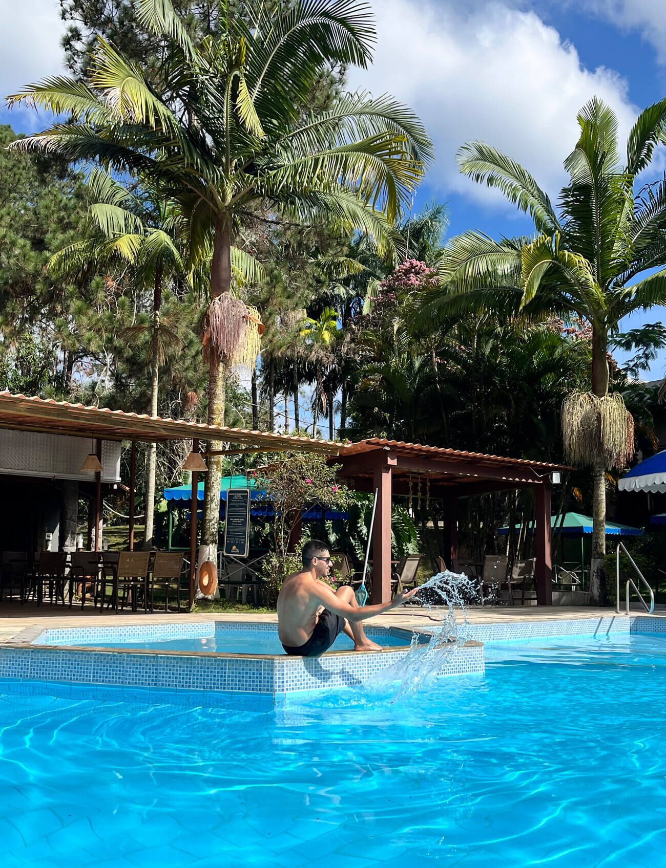 A man sitting by a pool with palm trees and a covered seating area in the background.