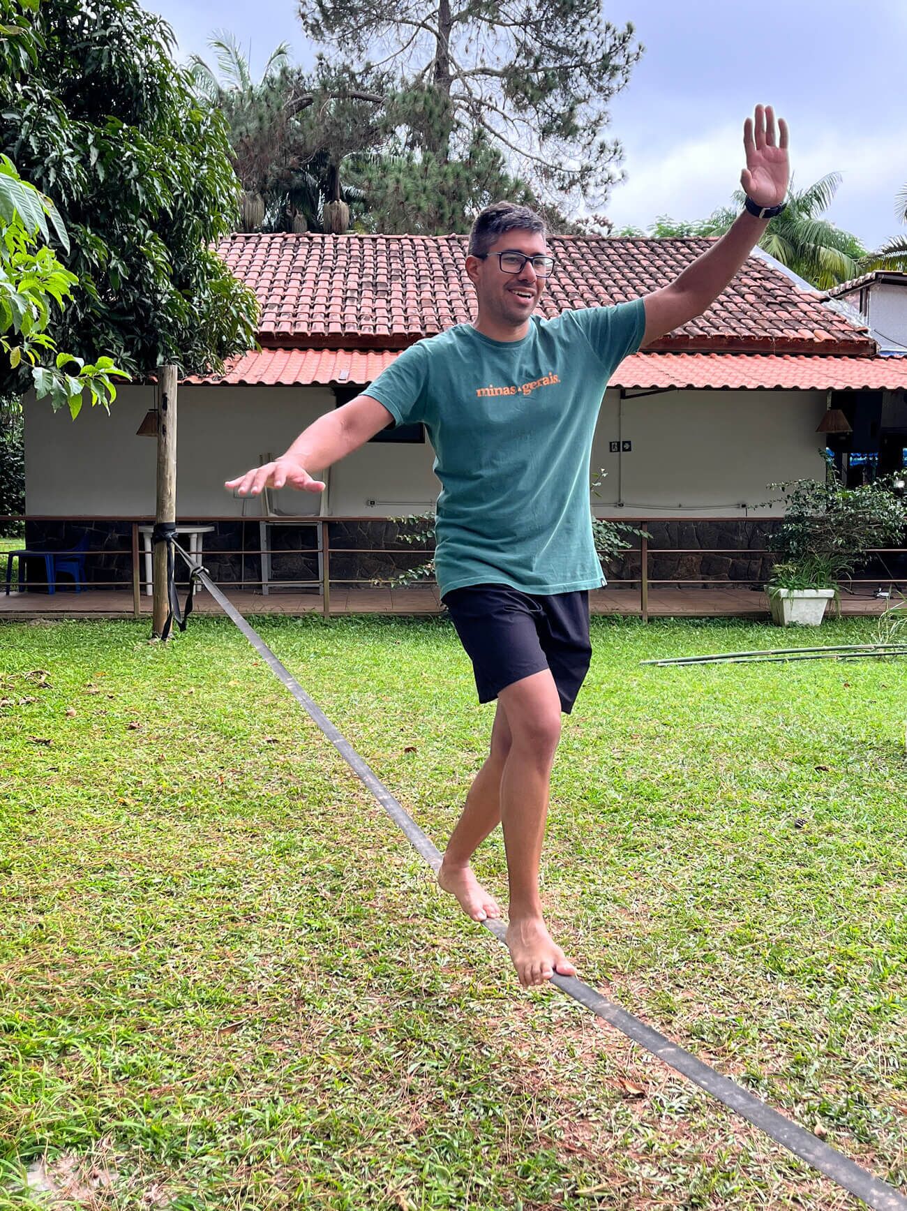 Uma pessoa equilibrando-se numa fita bamba em uma área gramada, com uma casa ao fundo.