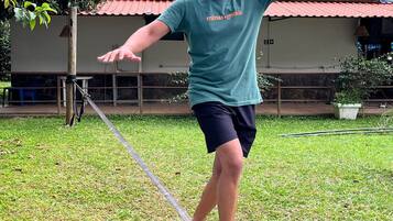 Uma pessoa equilibrando-se numa fita bamba em uma área gramada, com uma casa ao fundo.